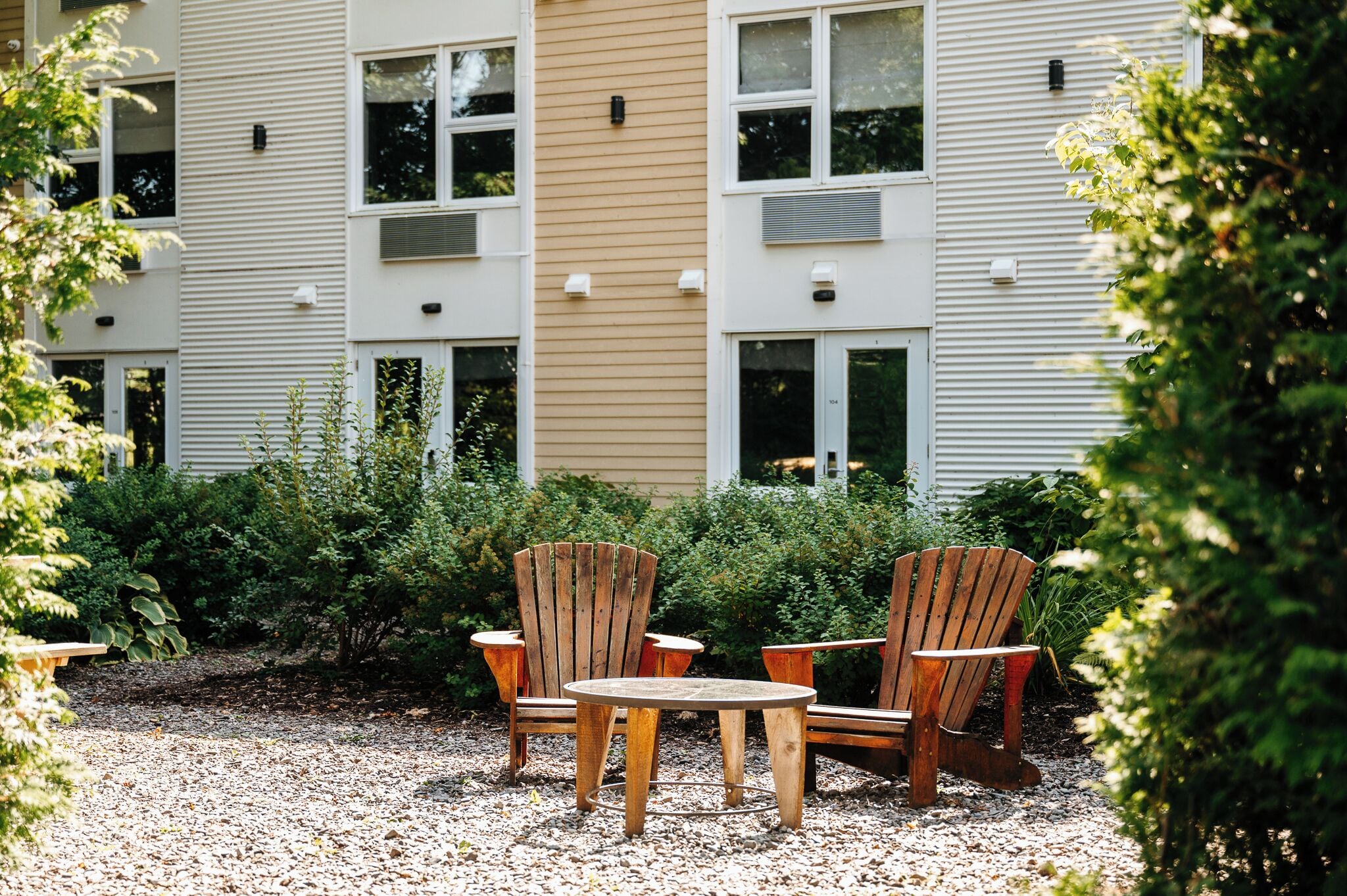 Un bâtiment à l’extérieur beige et blanc, avec une cour en gravier, des chaises et une table en bois.