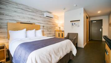 A hotel room with a large bed, a wooden headboard, an air conditioning unit, a lamp, a chair, a door, and a wall-mounted decoration.