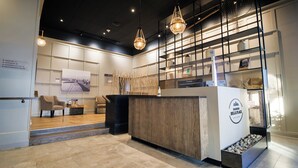 A modern reception area with a wooden reception desk, seating area with chairs, and a wall-mounted display with a framed picture and a ViewSonic monitor. - Manoir Belle Plage (Carleton-sur-Mer)