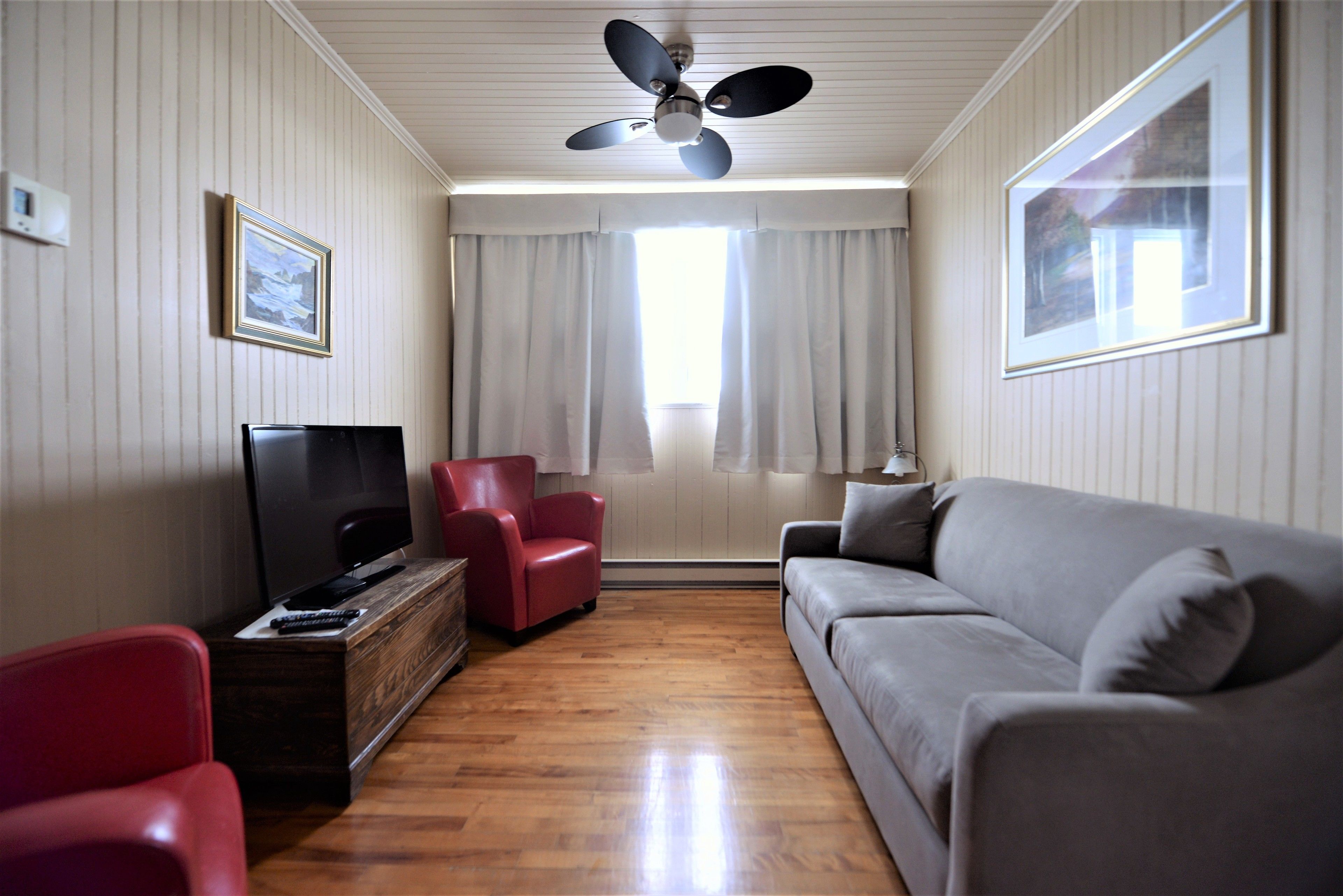 Una sala de estar con una televisión de pantalla plana, sofás rojos y grises, una mesa de centro de madera y un ventilador de techo.