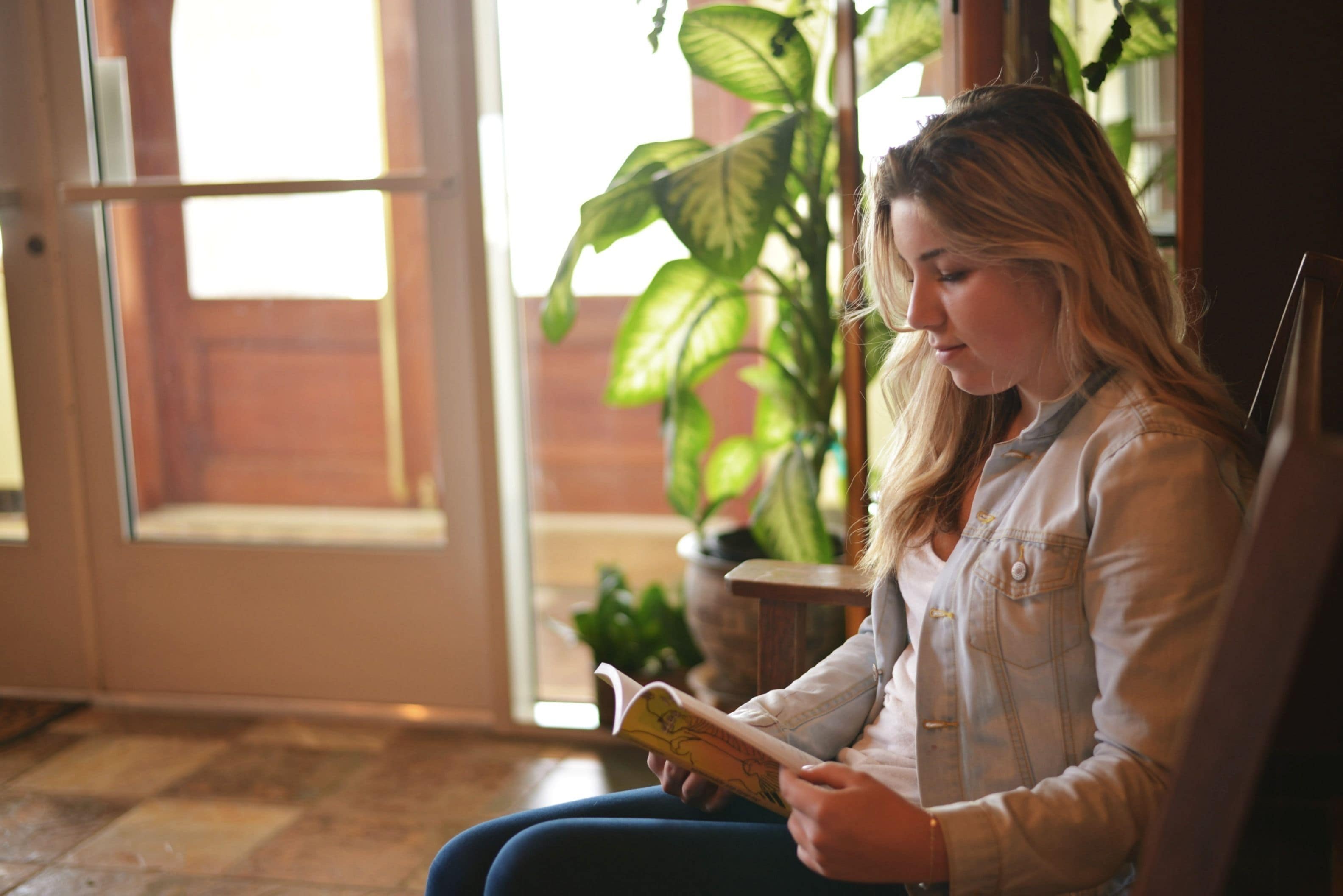 A person sitting indoors, reading a book, with a potted plant and a door in the background.