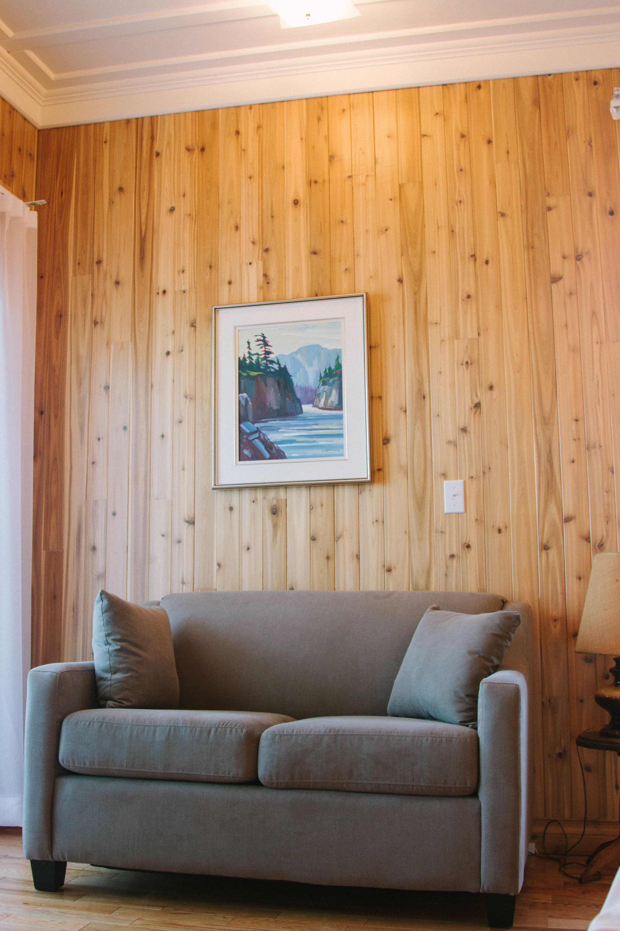 A room with a wooden paneled wall, a framed painting of a landscape, and a grey sofa with cushions.