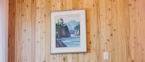 A room with a wooden paneled wall, a framed painting of a landscape, and a grey sofa with cushions.