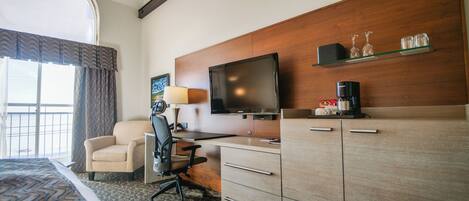 Una habitación de hotel con cama, escritorio y silla, televisor, cafetera y estante con vasos.
