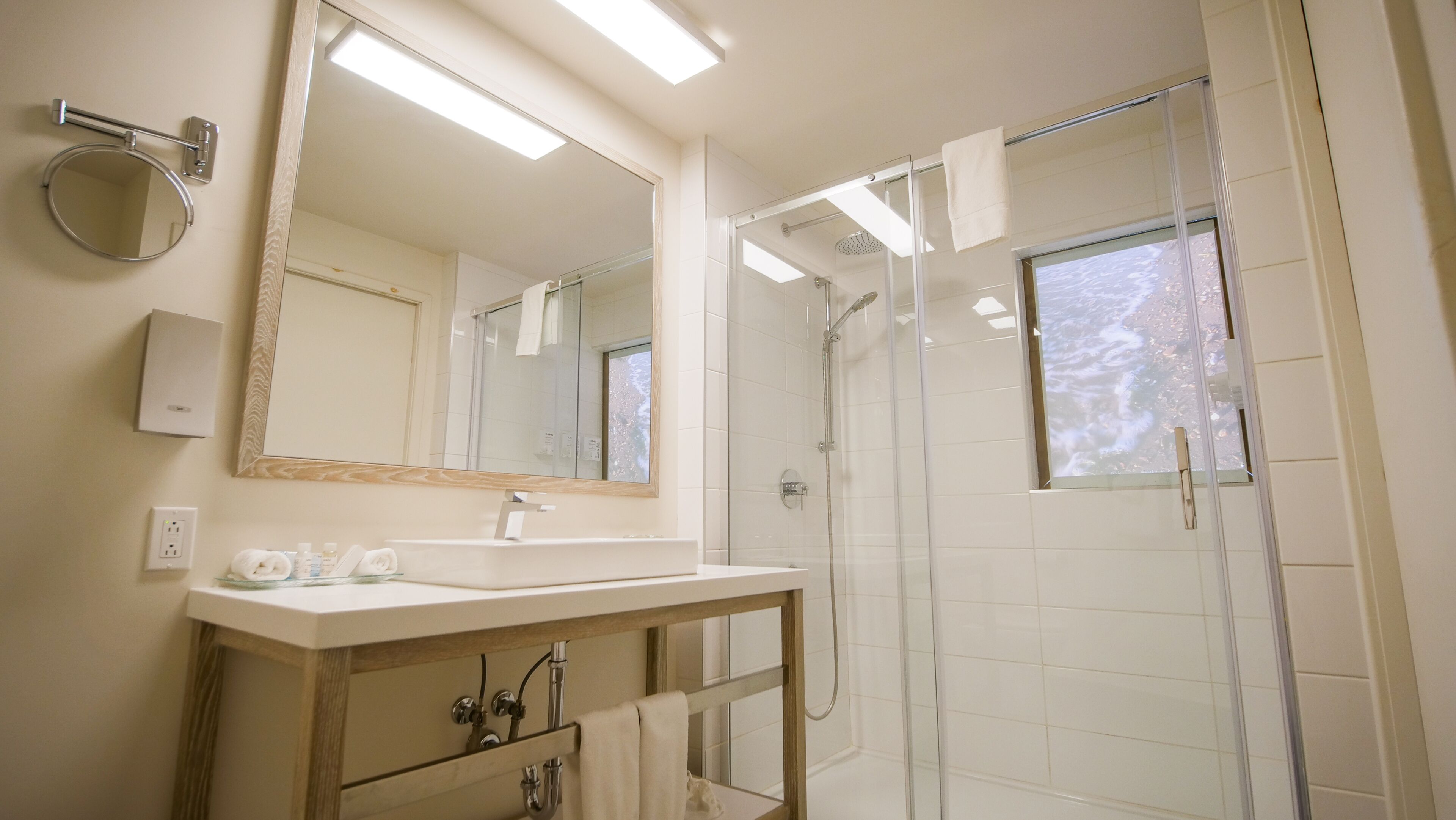 Un baño moderno con un espejo grande, un espejo redondo, un lavabo con tocador y una ducha con mampara de vidrio.