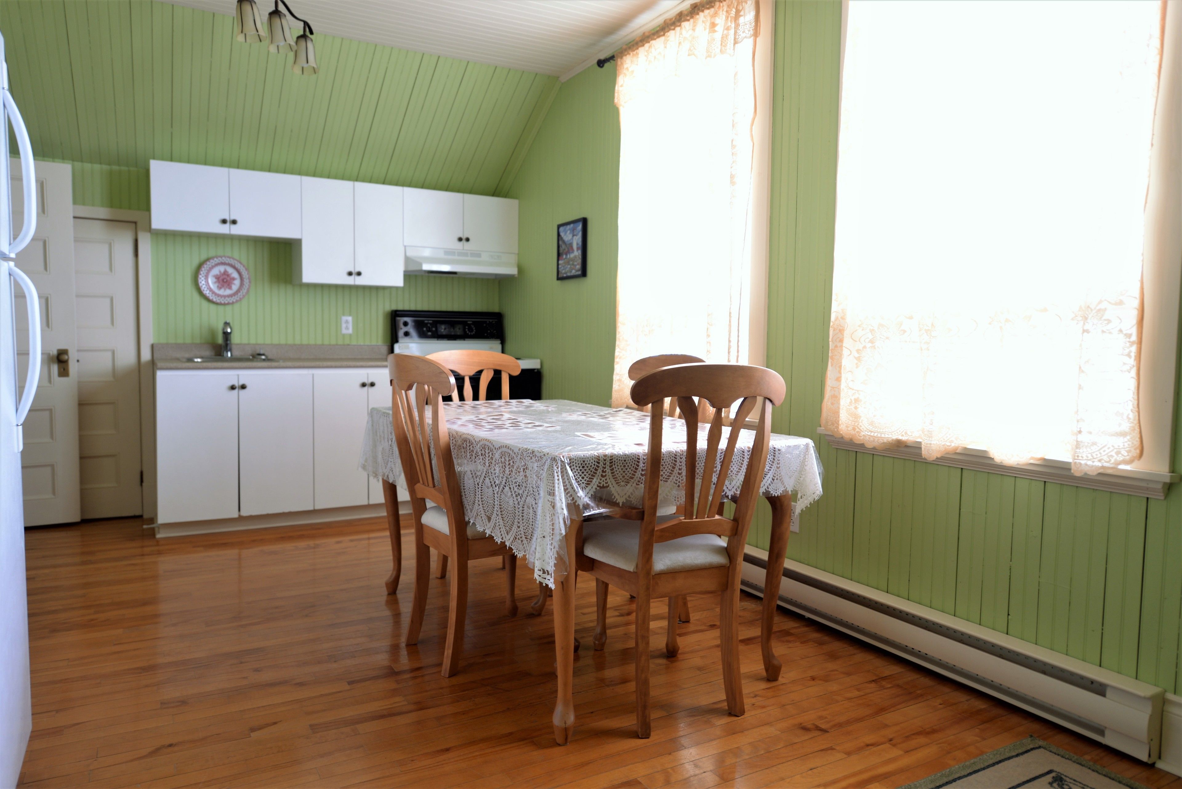Un comedor con una mesa dispuesta para cuatro personas, sillas de madera, un mantel de encaje blanco y una cocina al fondo.