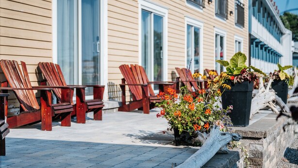 A row of wooden chairs and tables outside a building with large windows and a stone wall. - Manoir Belle Plage (Carleton-sur-Mer)