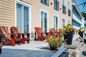 A row of wooden chairs and tables outside a building with large windows and a stone wall. - Manoir Belle Plage (Carleton-sur-Mer)