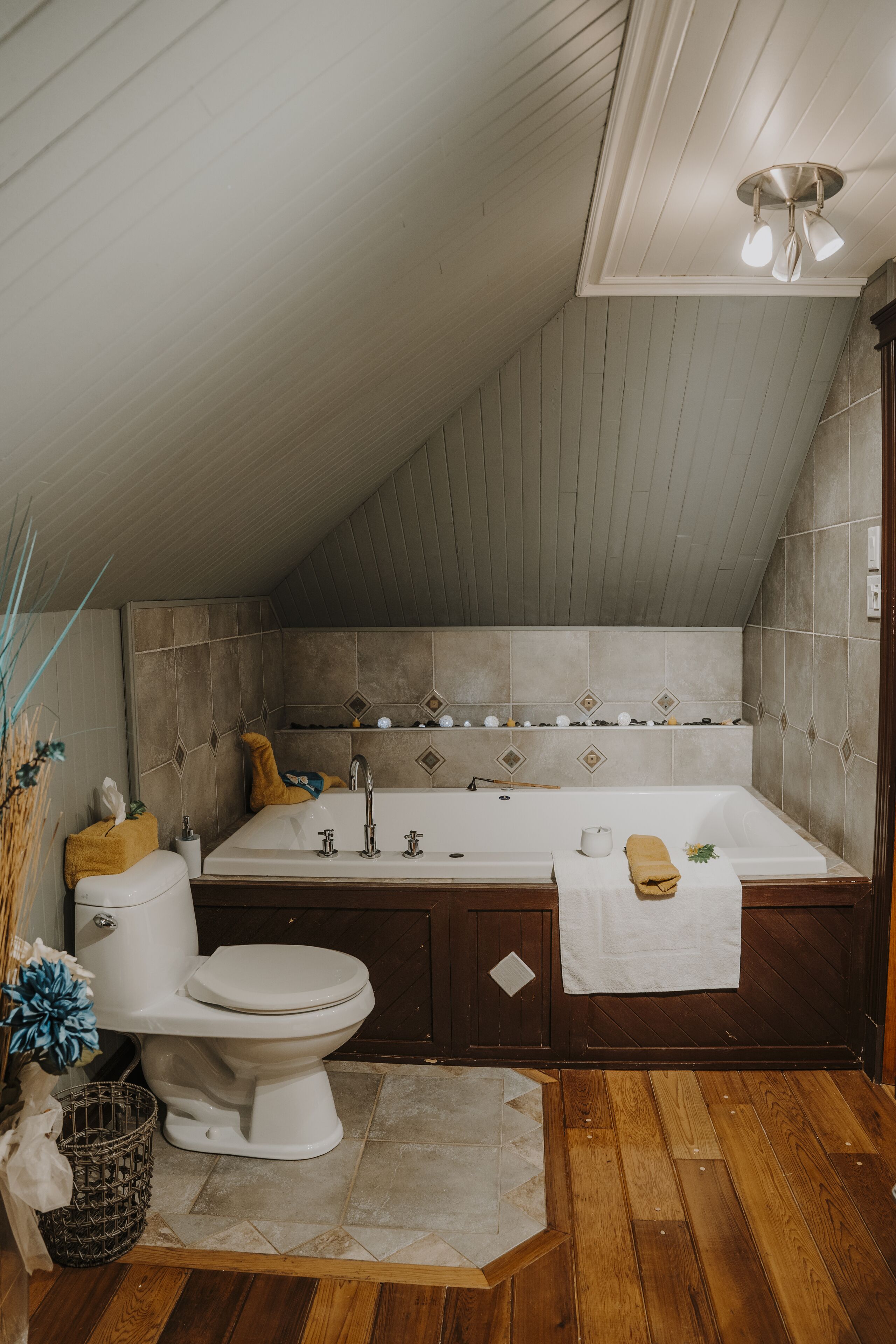 A bathroom with a toilet, sink, and bathtub, wooden flooring, and a sloped ceiling.