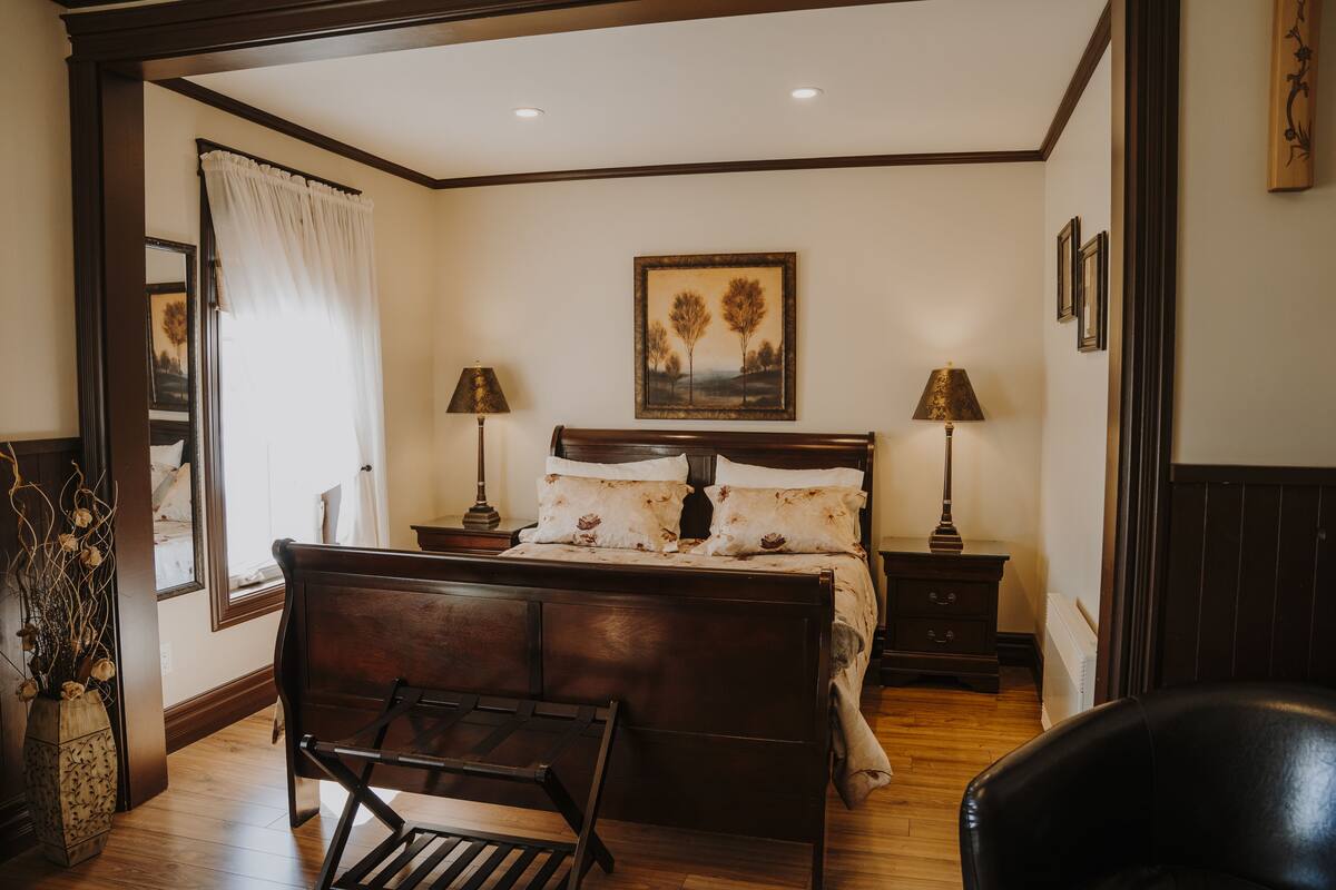 Quarto com cama de madeira, duas mesas de cabeceira com abajures, uma janela com cortinas e um quadro na parede.