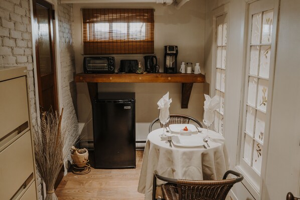 A compact kitchen with a refrigerator, a coffee maker, and a toaster. There is a dining table with chairs and a window with blinds.