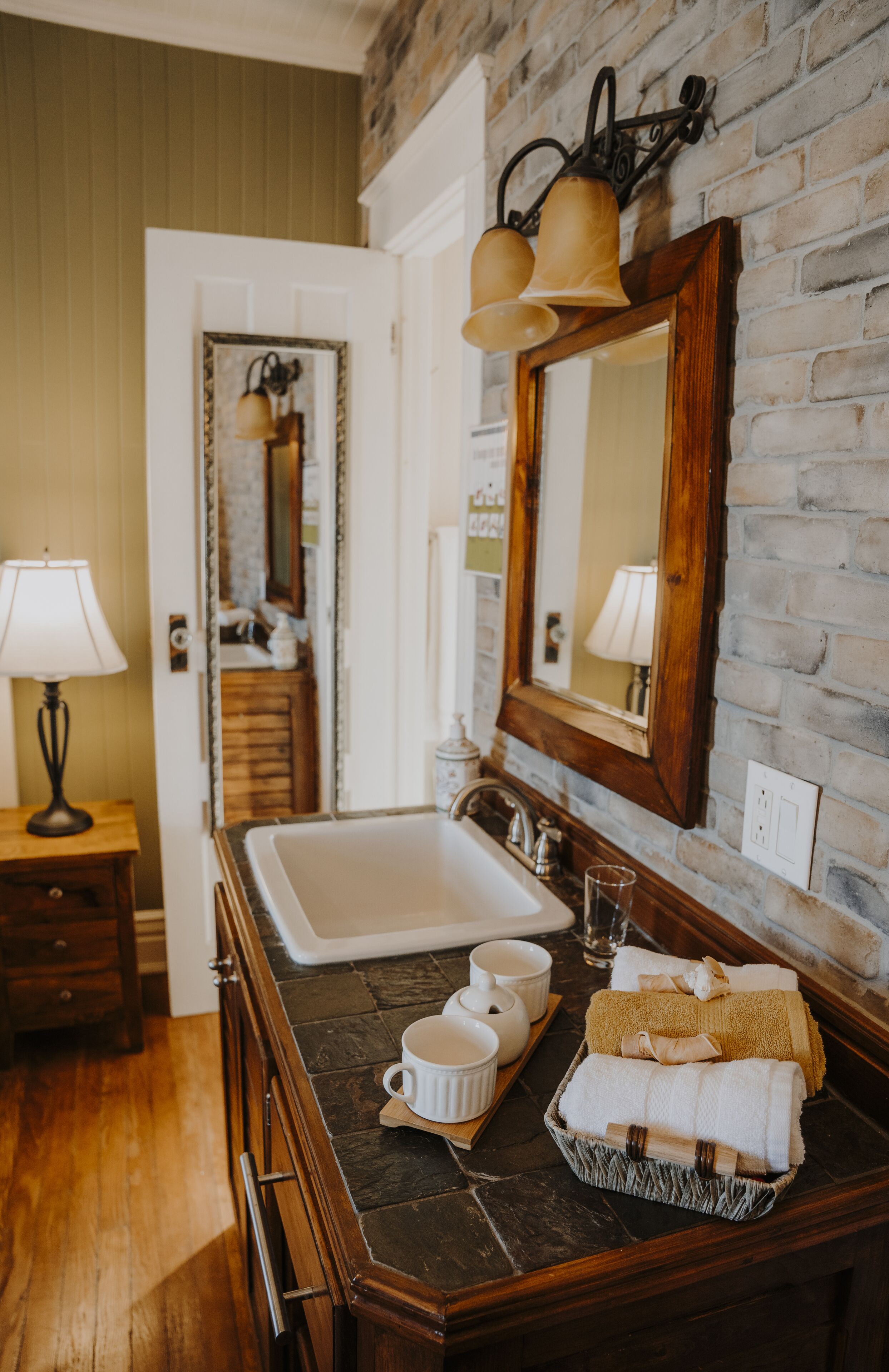 Une salle de bain avec un grand lavabo, un meuble-lavabo en bois et un miroir. On y trouve des serviettes et des tasses sur le meuble-lavabo.