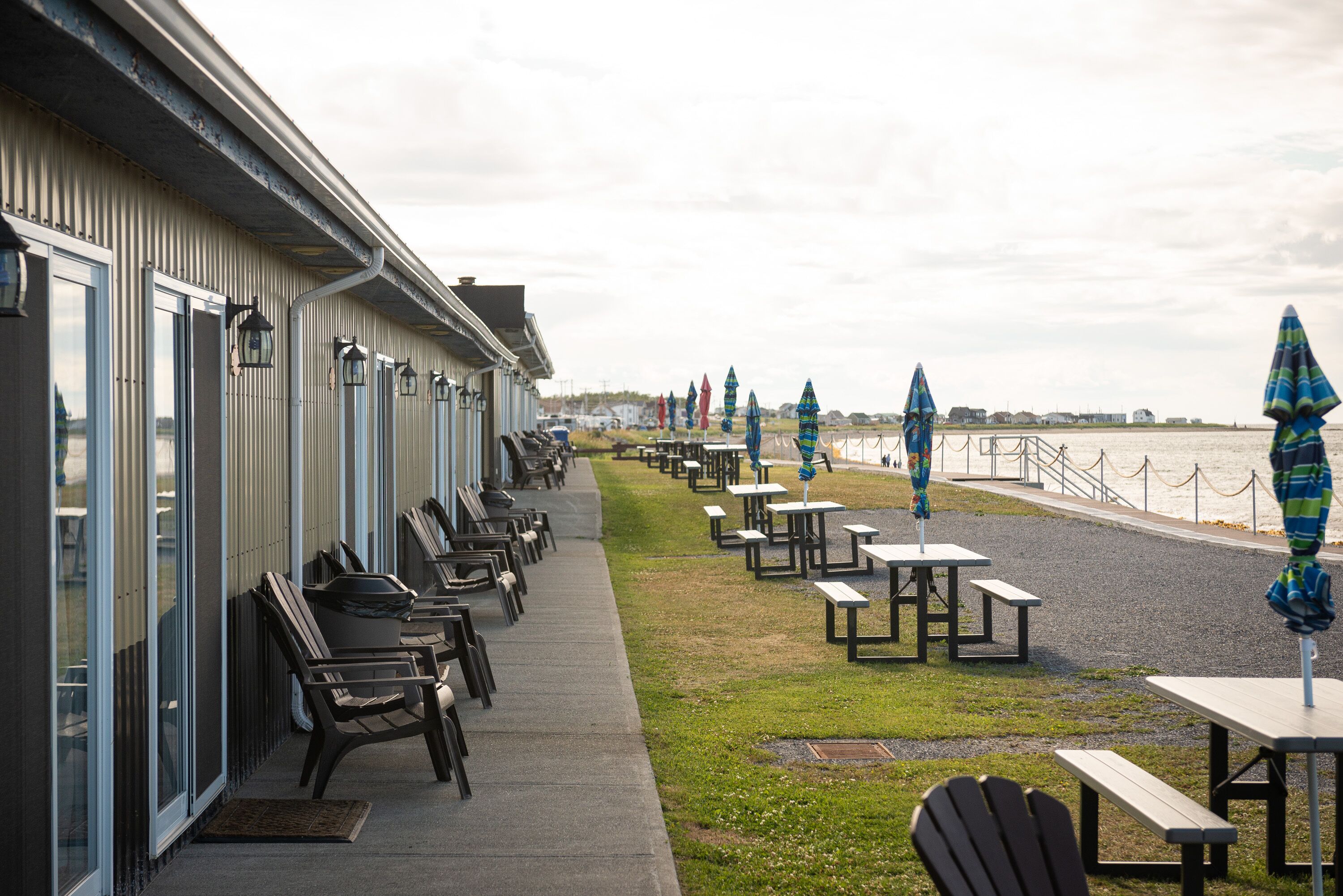 A row of outdoor seating with tables and chairs along a building's side, overlooking a body of water.