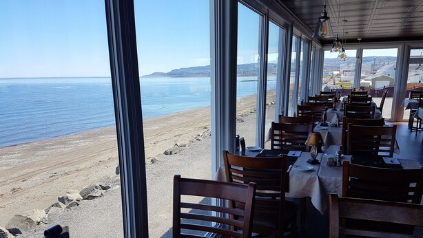 A seaside restaurant with large windows offering ocean views, wooden tables and chairs, and a menu sign. - Manoir sur Mer (Sainte-Anne-des-Monts)