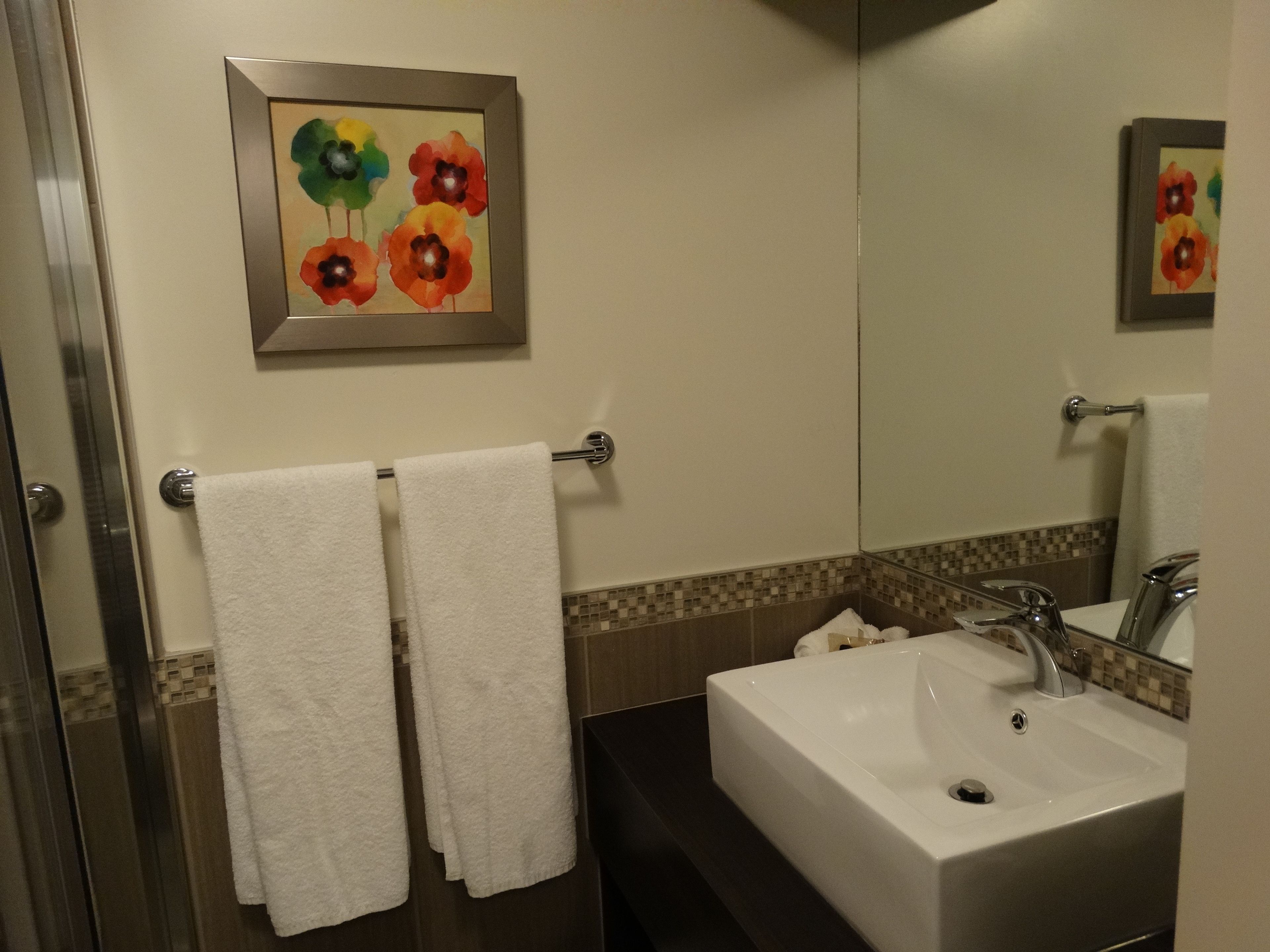 Une salle de bain avec un lavabo blanc, un miroir et un porte-serviettes sur lequel sont posées deux serviettes blanches. Un tableau encadré représentant des fleurs est accroché au mur.