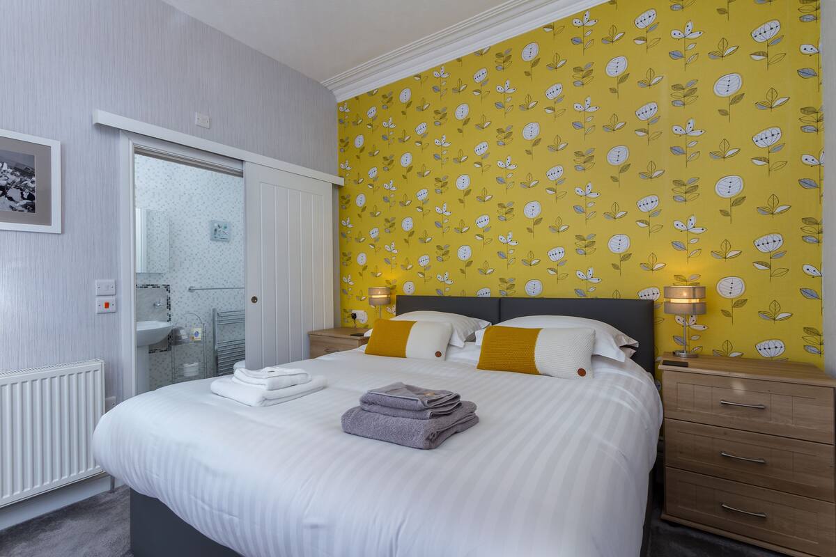 Una cama bien hecha con sábanas blancas y una toalla doblada, una mesita de noche con una lámpara, y una pared con estampado floral.