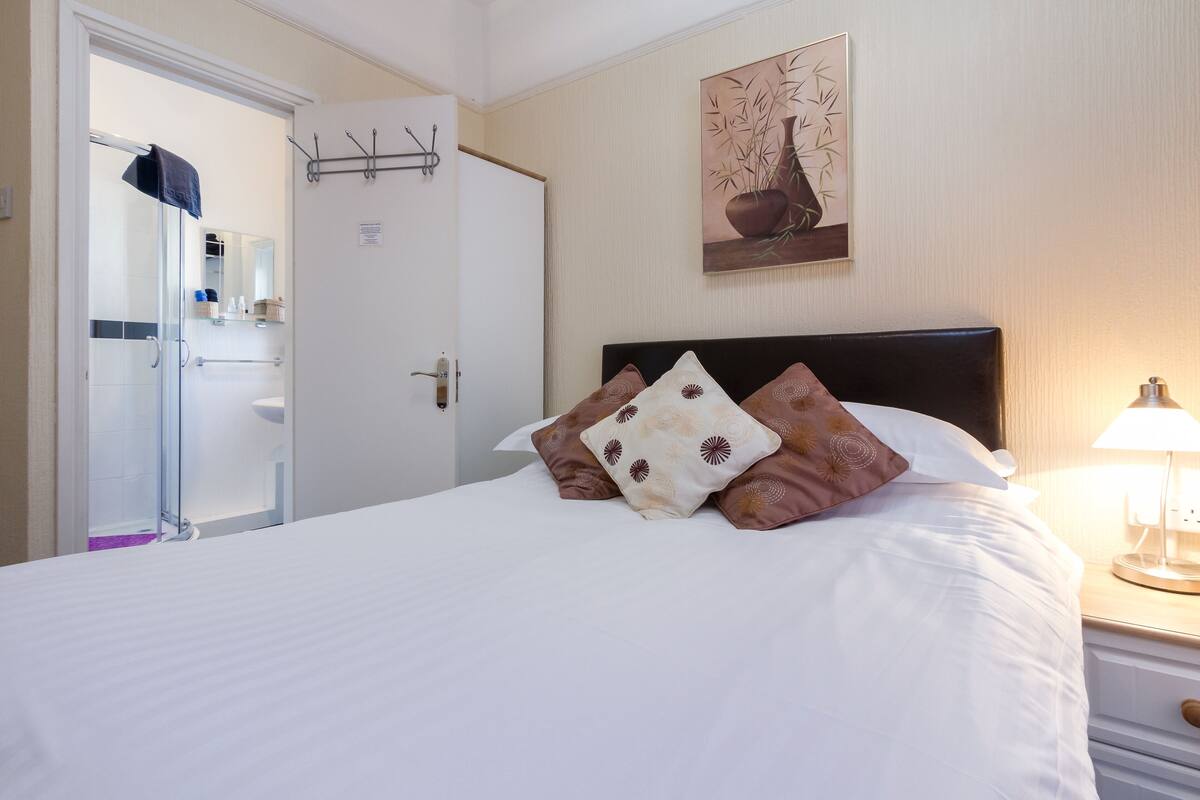 Una habitación de hotel con cama, mesita de noche, lámpara, baño con cabina de ducha de vidrio y una obra de arte colgada en la pared.