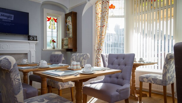 Une salle à manger avec une table en bois dressée pour quatre personnes, entourée de chaises rembourrées. Un téléviseur est fixé au mur, au-dessus de la cheminée.