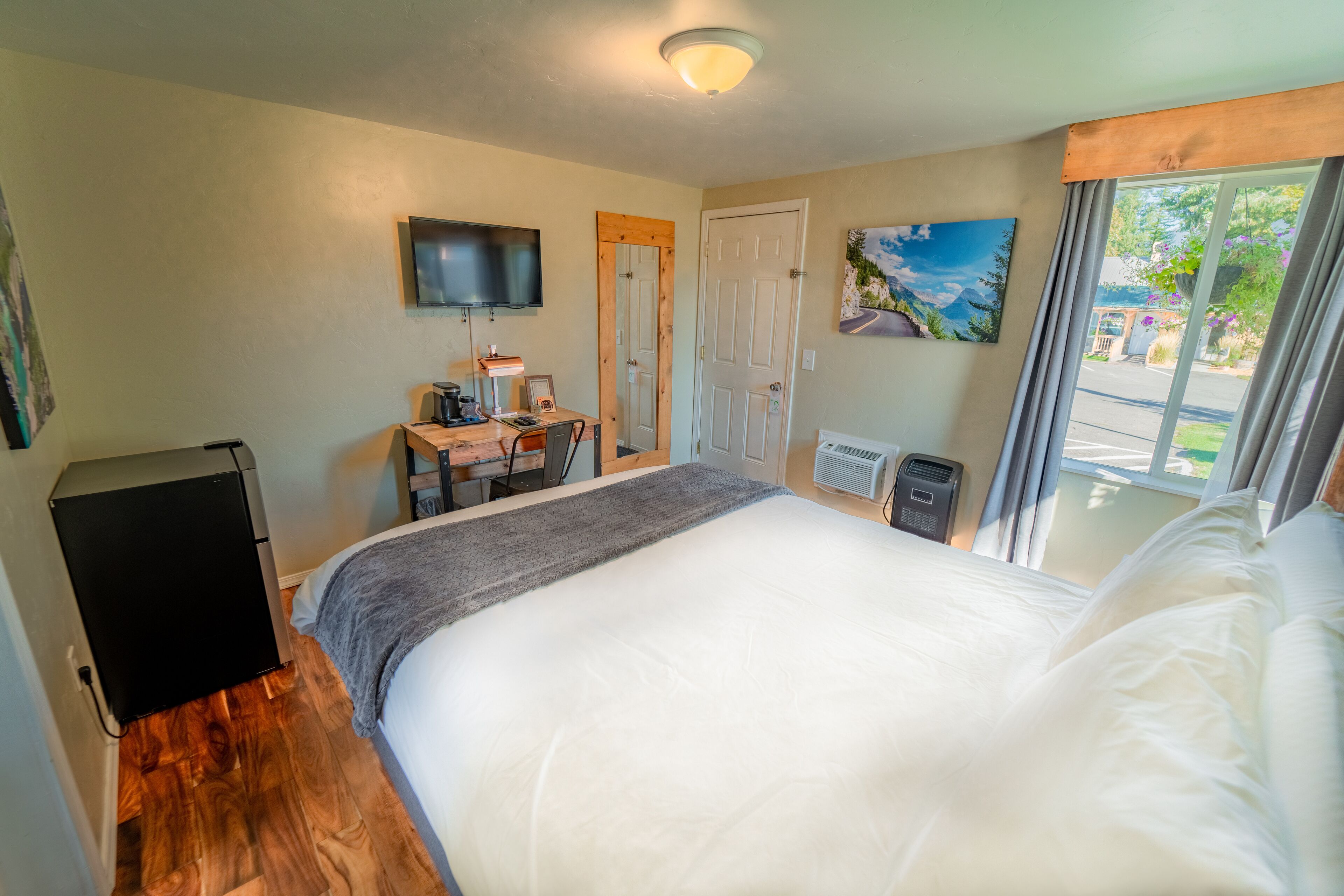 Ein Schlafzimmer mit einem großen Bett, einem Schreibtisch mit Computer, einem an der Wand befestigten Fernseher und einem Fenster mit Vorhängen.