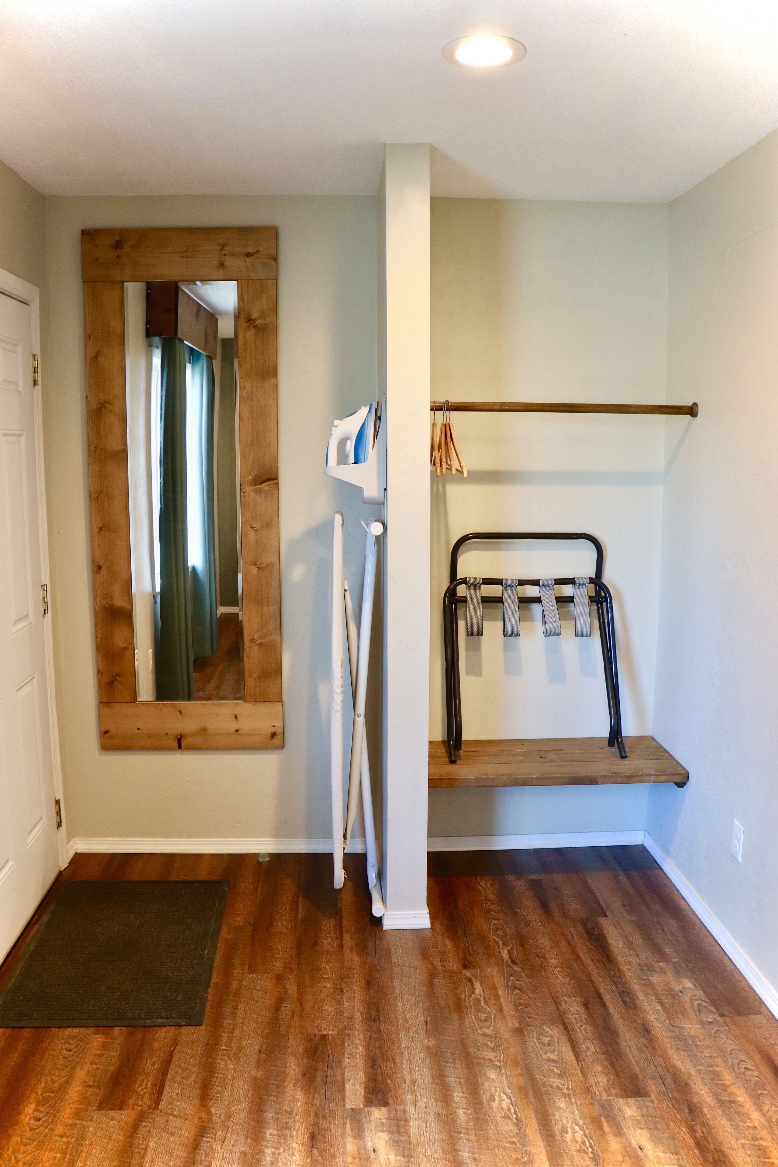 Una habitación con suelo de madera, un espejo con marco de madera, una puerta blanca y un estante con toallero.