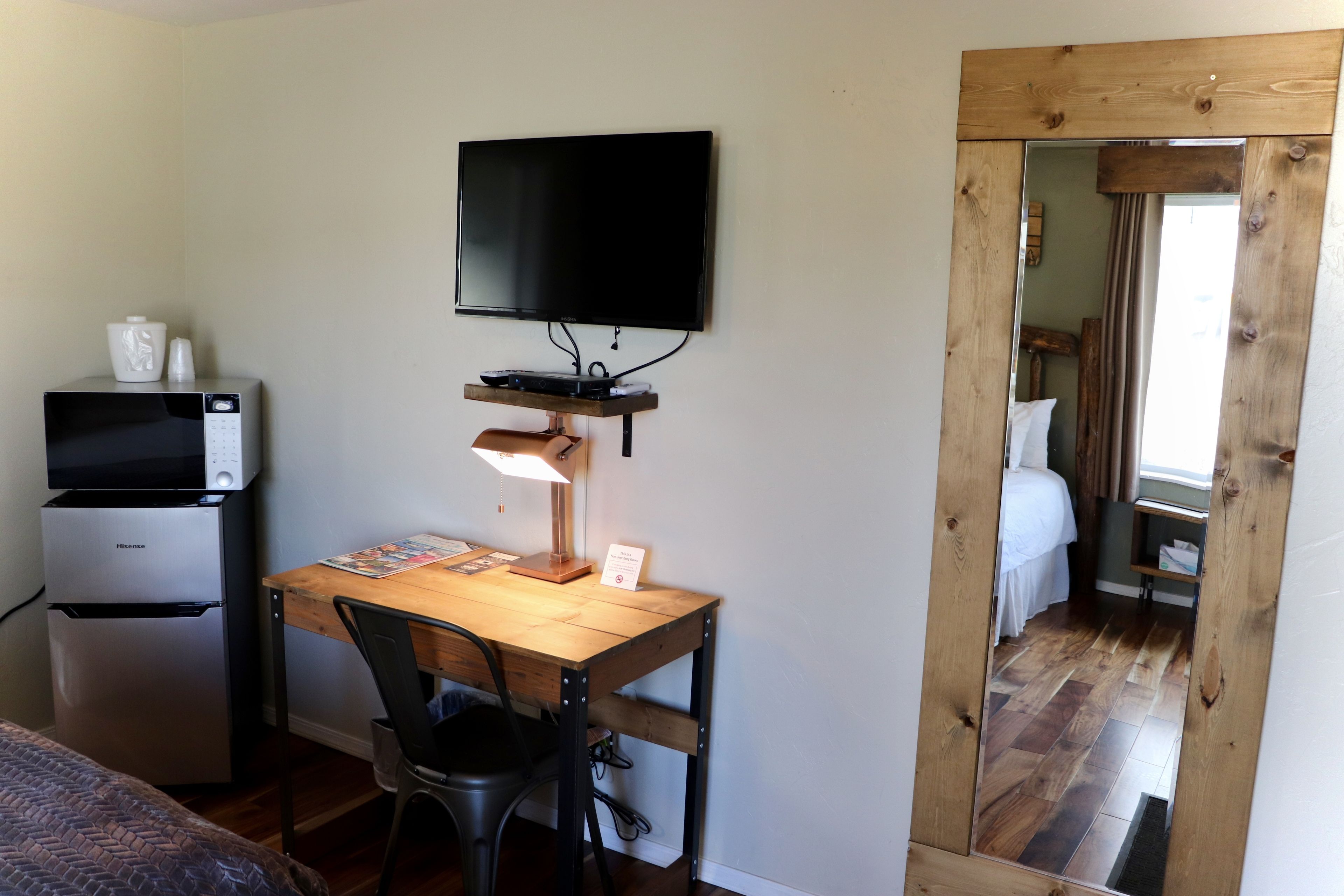 A compact room with a bed, a desk with a computer and a flat-screen TV mounted on the wall, a refrigerator, and a wooden-framed mirror.