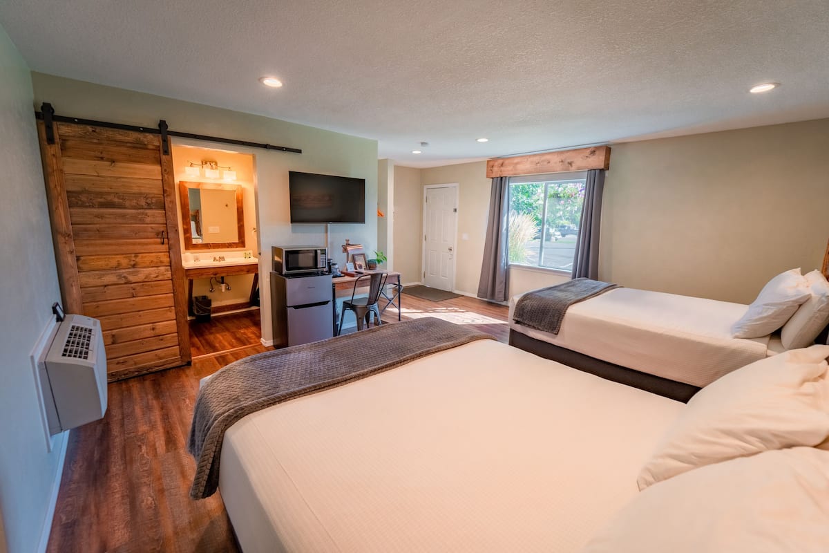 A hotel room with two beds, a desk with a computer and a flat-screen TV, a sliding door closet, and a large window with a view of greenery.