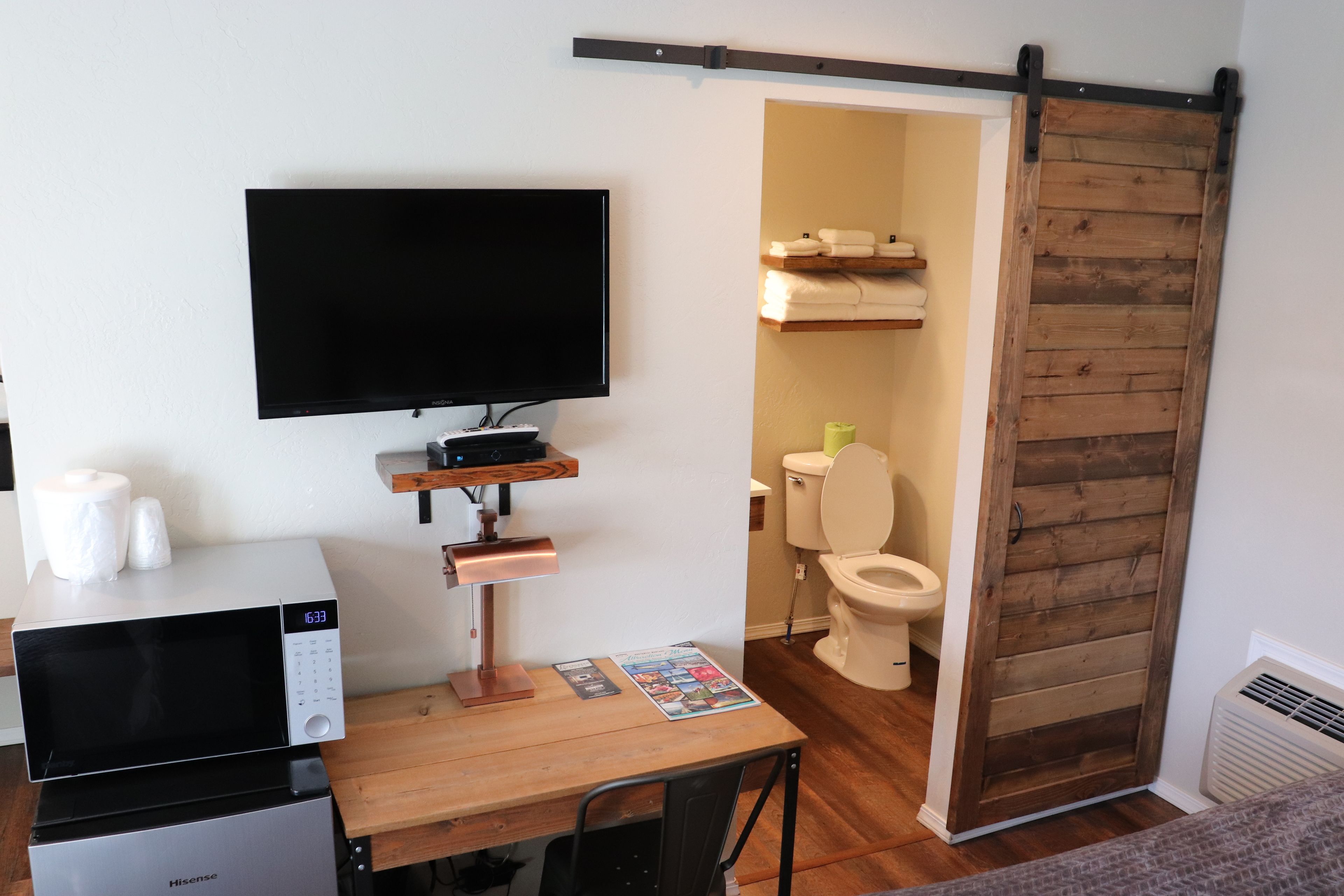 Habitación compacta con televisor de pantalla plana, microondas, una pequeña mesa con una revista y una puerta corredera que da al baño.