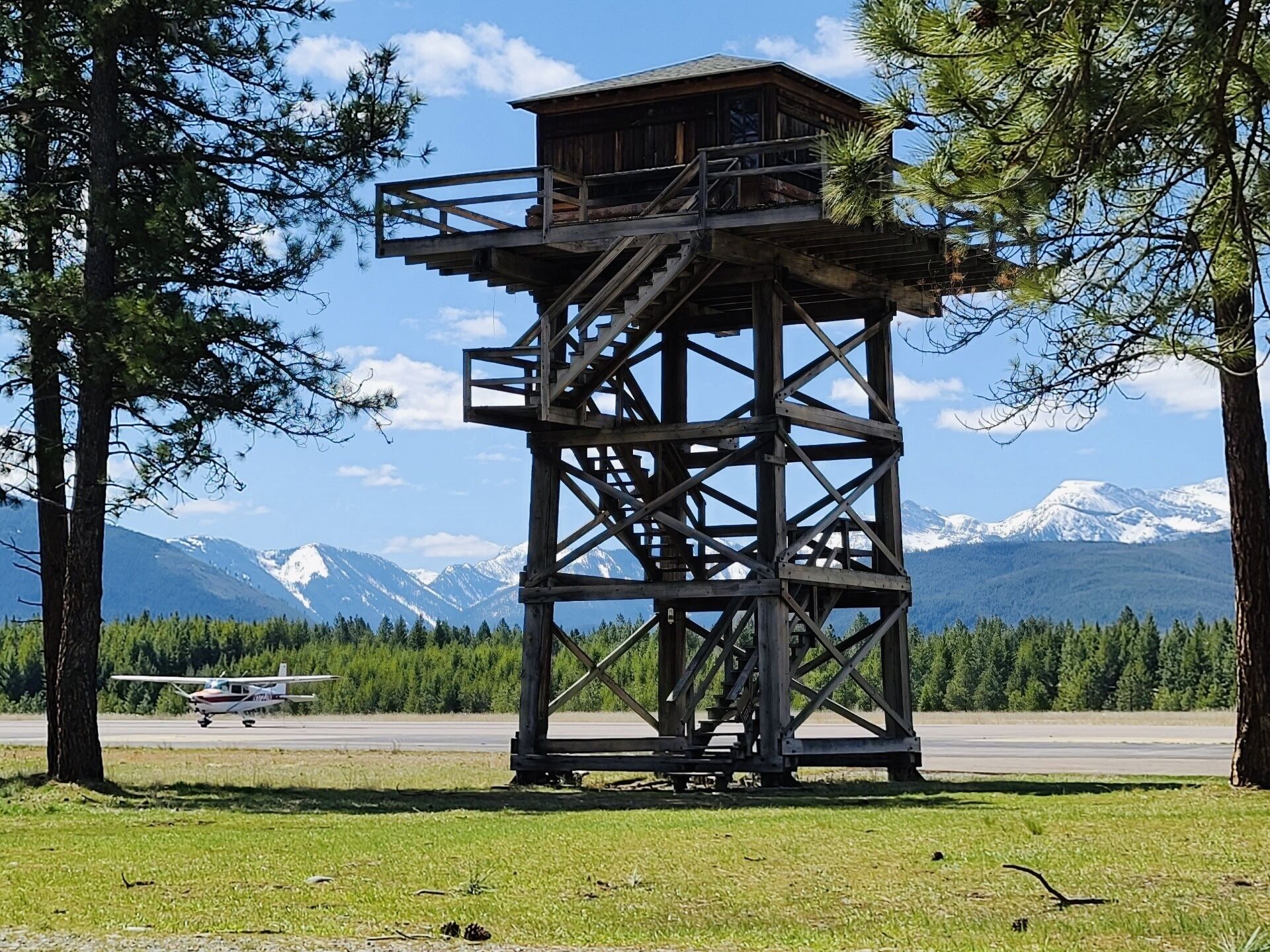 A wooden observation tower with a staircase, surrounded by trees and a backdrop of snow-capped mountains.