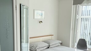 A neatly arranged bedroom with a bed, a bedside table, a chair, a window, and a wall-mounted fan. - Westward Bed & Breakfast (Newquay)