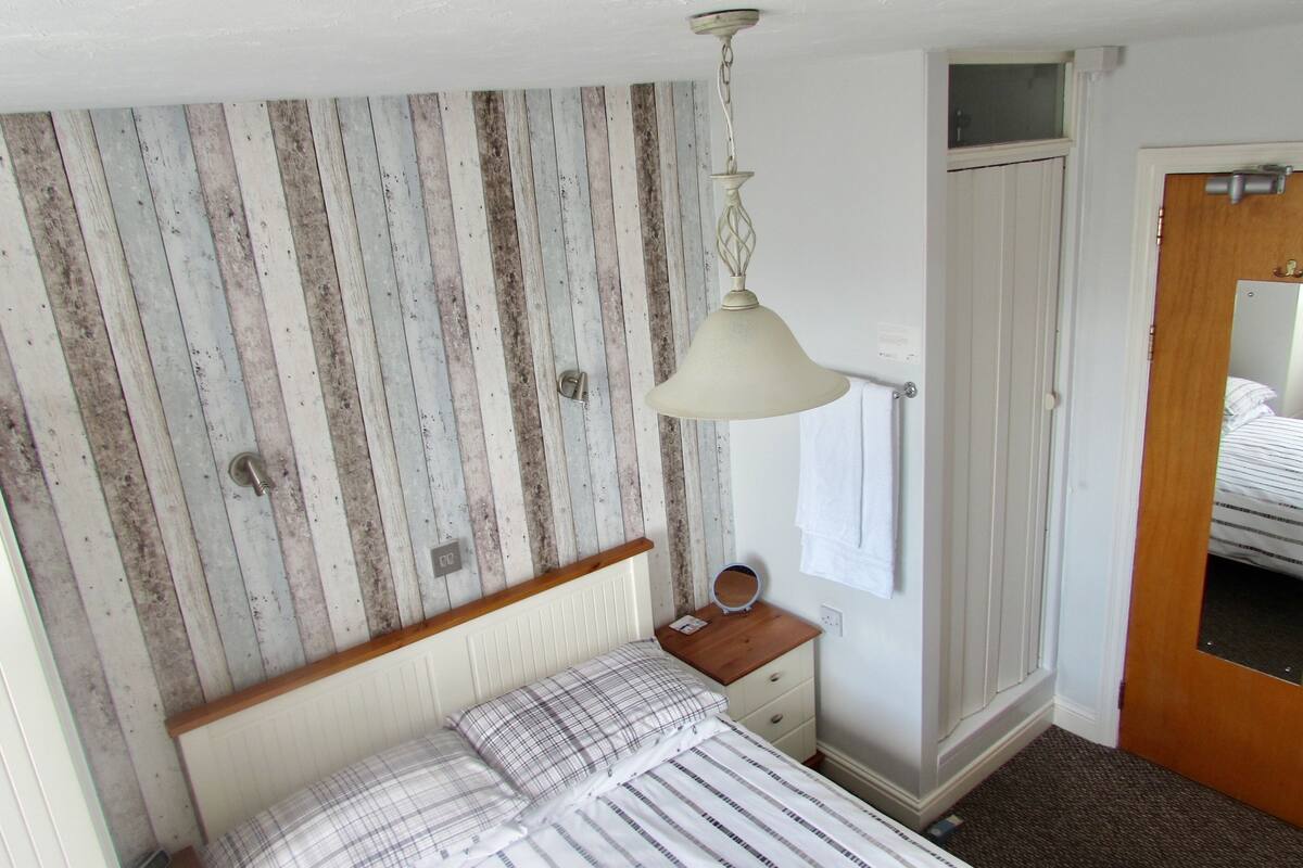 A bedroom with a wooden wall panel, a bed with striped bedding, a bedside table with a mirror, and a door with a mirror.
