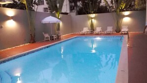 A rectangular swimming pool with a tiled edge, surrounded by a low wall. There are several white lounge chairs and a white umbrella near the pool. - Aparta Hotel Turey (Santo Domingo)