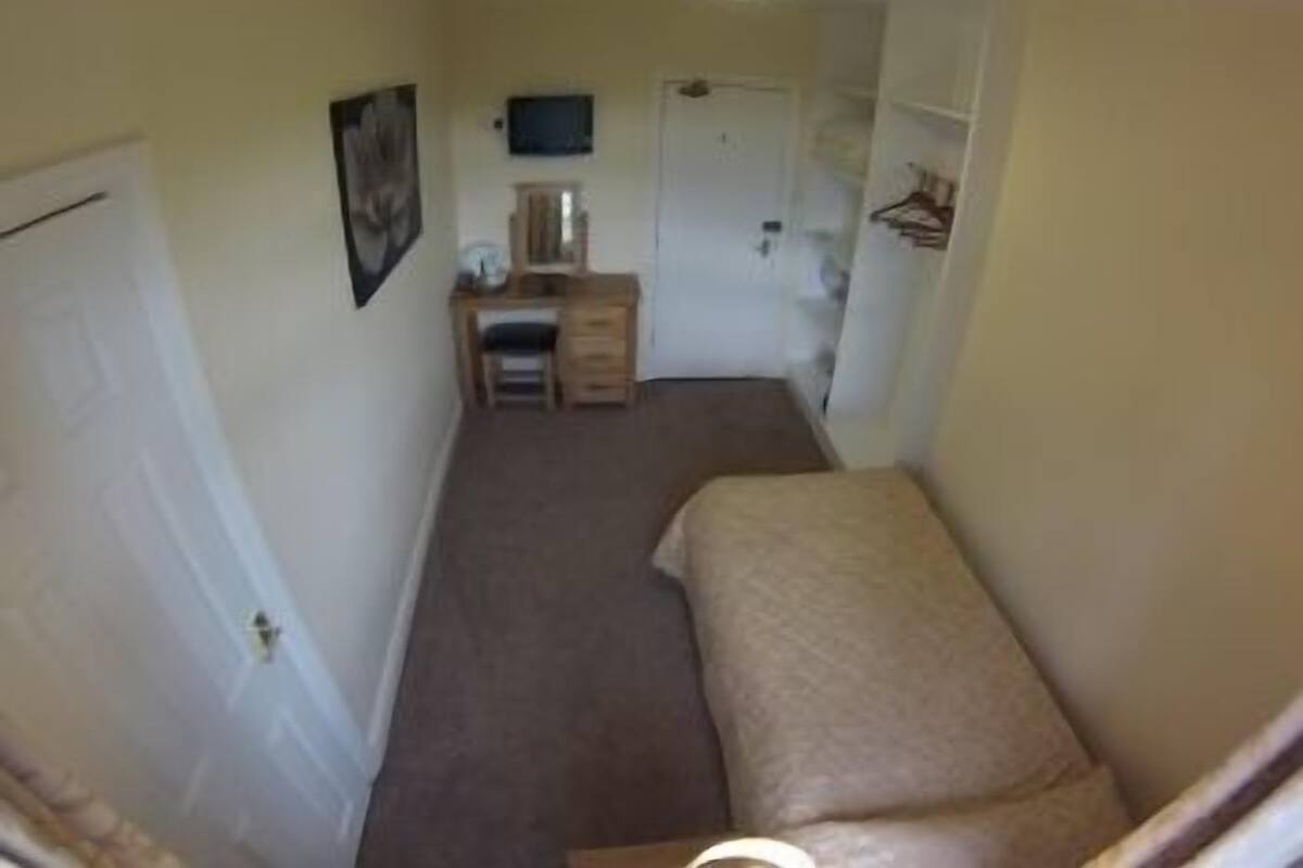 A narrow hallway with a bed on the right, a desk with a chair, and a door at the end.