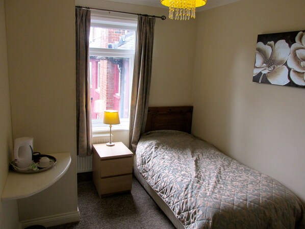 Un dormitorio con una cama individual, una mesita de noche con lámpara, un radiador y una ventana con cortinas.