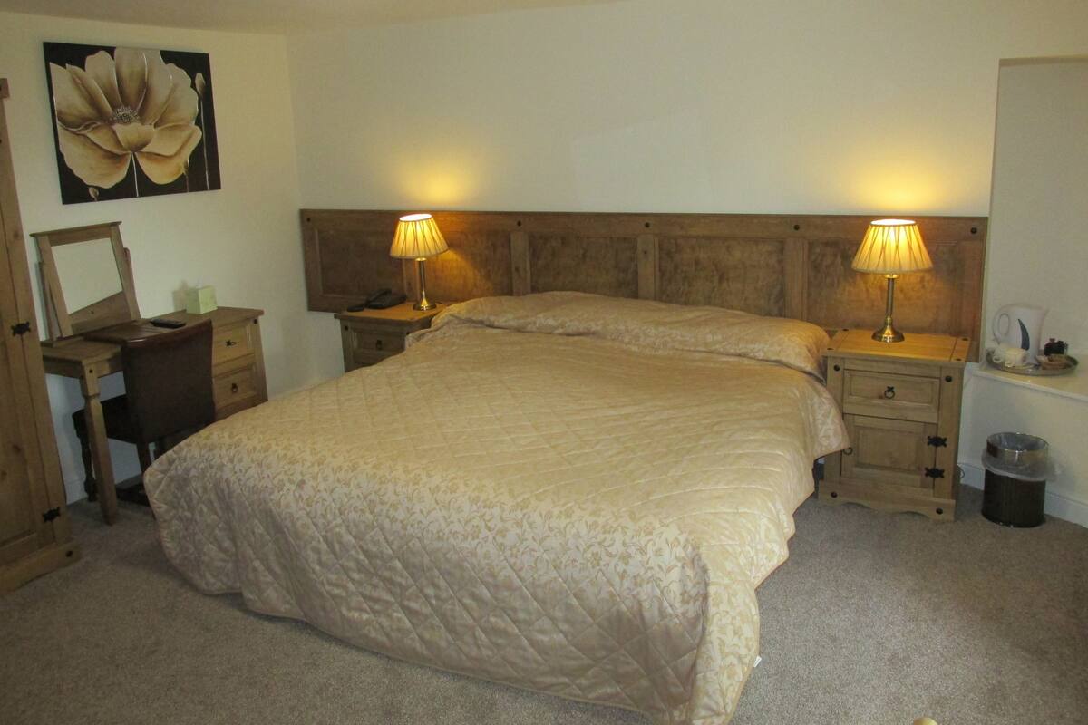 A bedroom with a large bed, two bedside tables with lamps, a wooden headboard, a desk with a chair, a mirror, and a wall-mounted artwork.