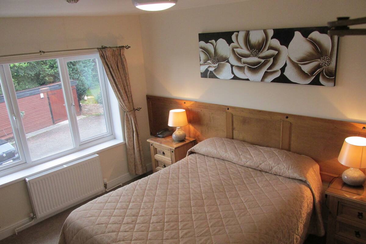 A bedroom with a bed, bedside lamps, a radiator, a window with curtains, and a floral painting on the wall.