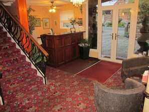 A hotel lobby with a wooden reception desk, a staircase with floral carpet, and a view of the outside through a glass door. - Edenhall Country House Hotel (Penrith)
