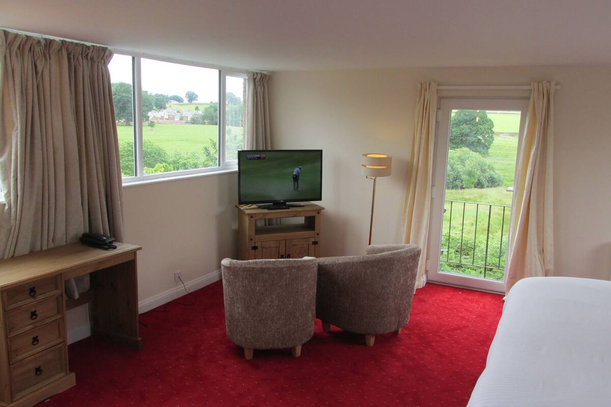 A hotel room with a bed, a desk with a telephone, two armchairs, a television on a wooden stand, and a balcony with curtains.
