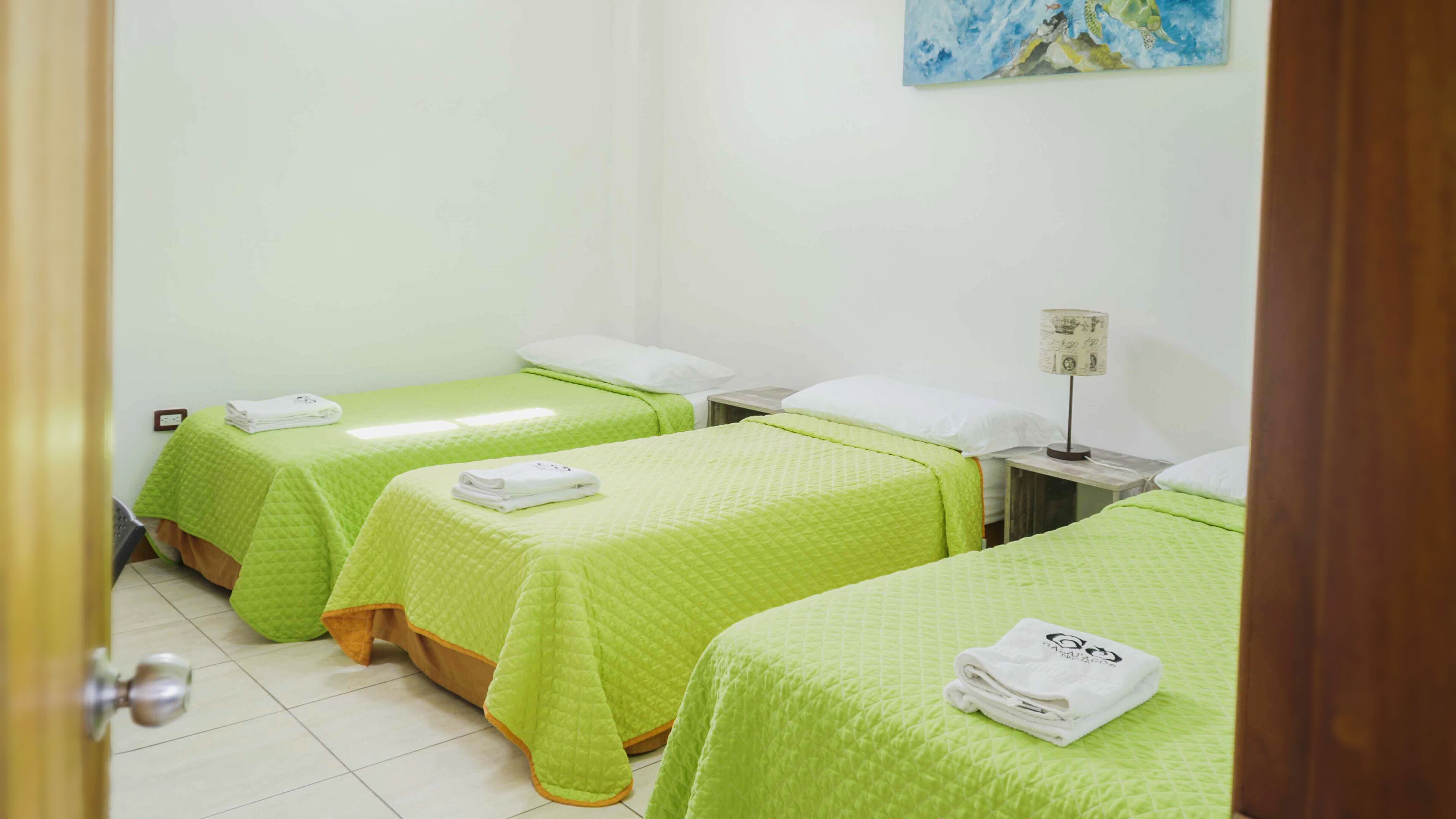 A hotel room with three beds, each with a green bedspread and white pillows. There is a small table with a lamp and a painting on the wall.