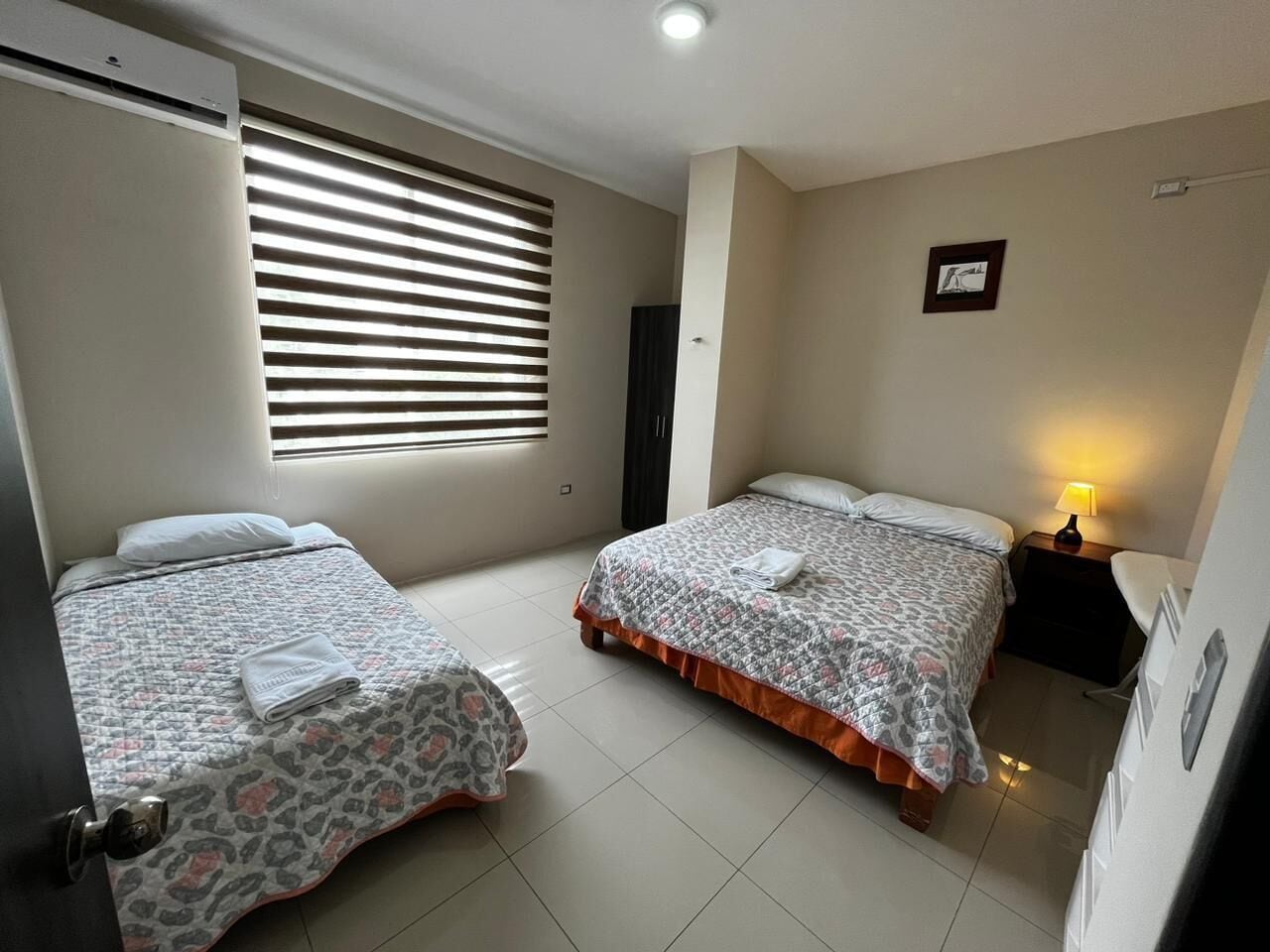 Habitación de hotel con dos camas, una ventana con persianas, una mesita de noche con lámpara y un cuadro enmarcado en la pared.