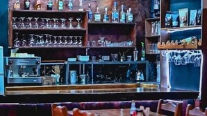 Zona de bar con estantes llenos de botellas y vasos, un juke box y sillas de madera.
