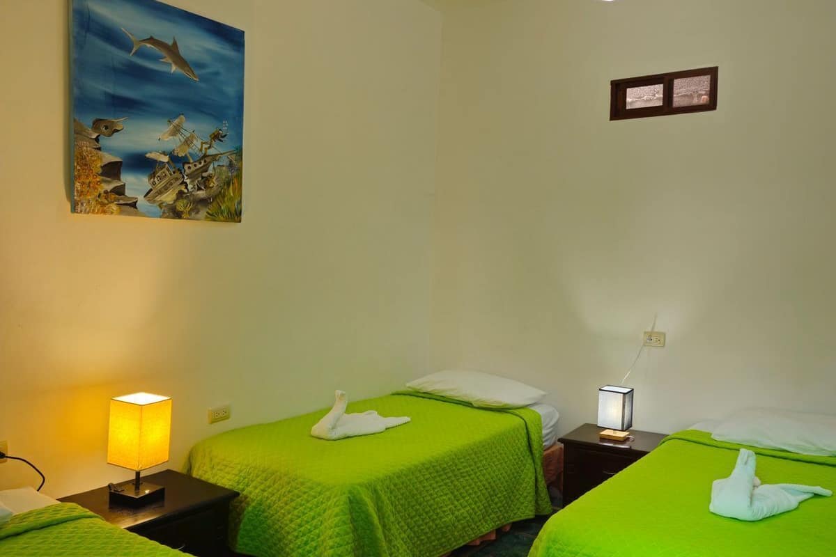 Ett hotellrum med två sängar, var och en med ett grönt överkast och en vit handduk formad som en svan. På väggen hänger en målning som föreställer ett marint motiv, till vänster står en lampa och till höger ett litet bord med en lampa.