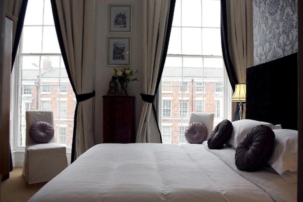 A hotel room with a large bed, two chairs, a nightstand, and a view of buildings through the window.