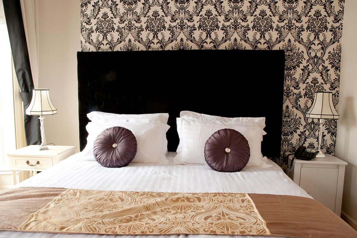 A hotel room with a large bed, two white pillows, two dark purple pillows, a decorative throw, a black headboard, patterned wallpaper, and bedside lamps.