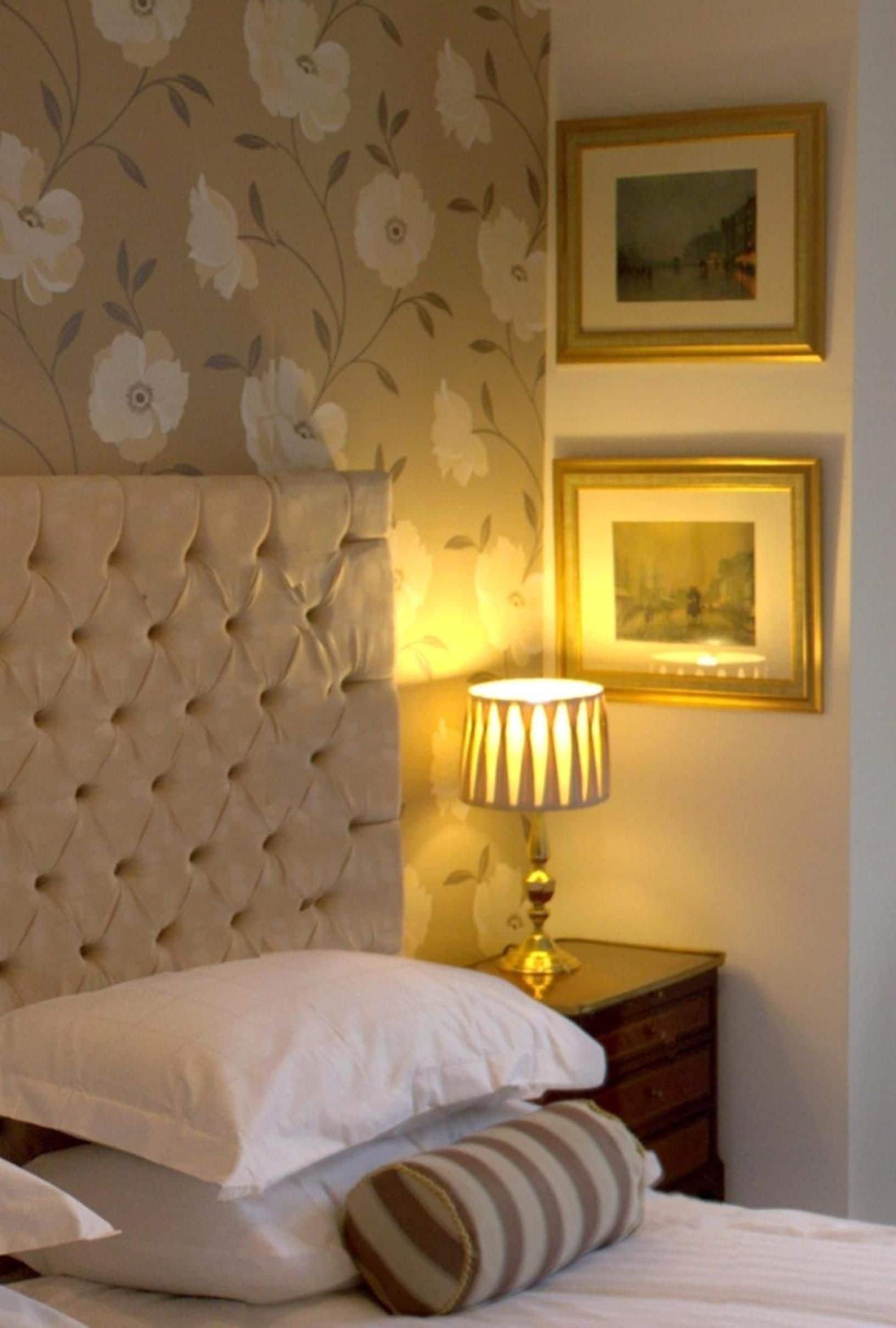 Habitación de hotel con cabecera capitonada, cama con ropa blanca, almohada a rayas, lámpara de noche y dos cuadros en la pared.
