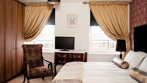 Habitación de hotel con una cama grande, televisión, armario de madera, sillón estampado y escritorio con cajones.