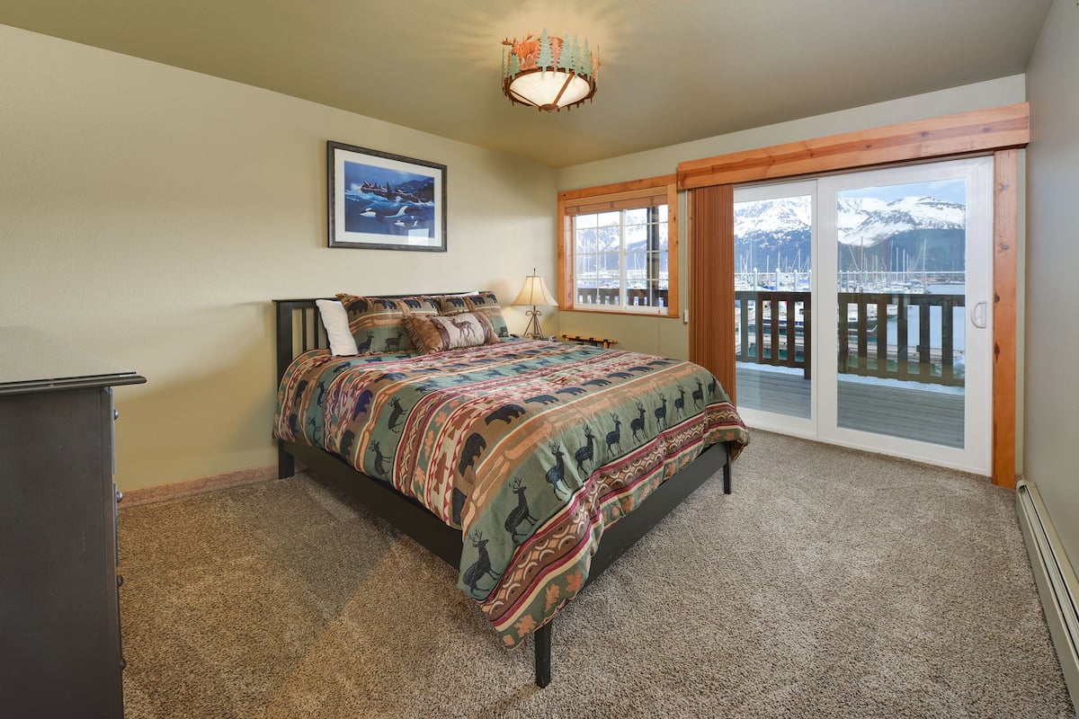 Ein Schlafzimmer mit Bett, Schminktisch, Fenster mit Blick auf eine Anlegestelle und Berge sowie einer Tür zu einem Balkon.