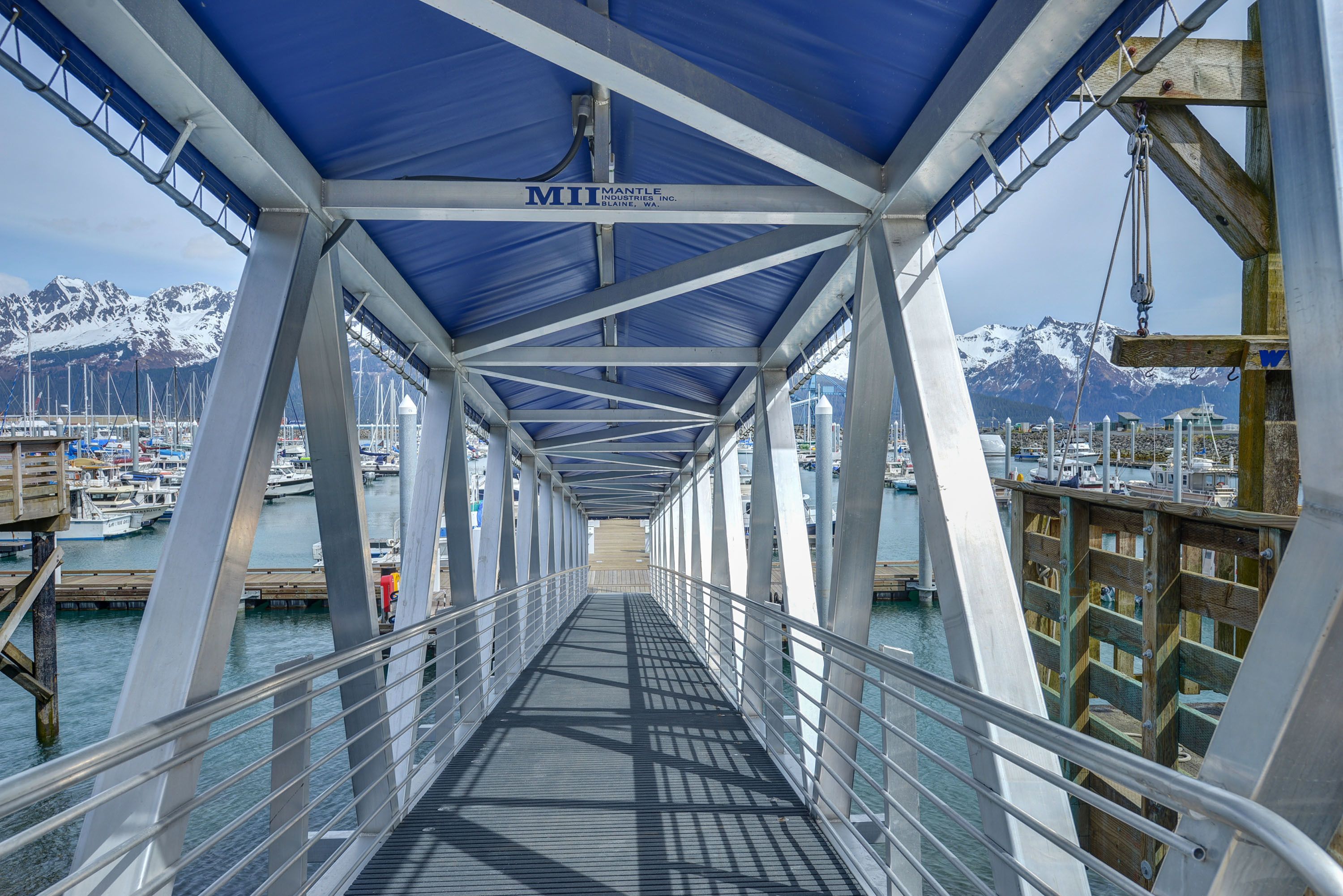 A covered pedestrian bridge with metal railings and a blue roof, overlooking a marina with boats and snow-capped mountains in the background.