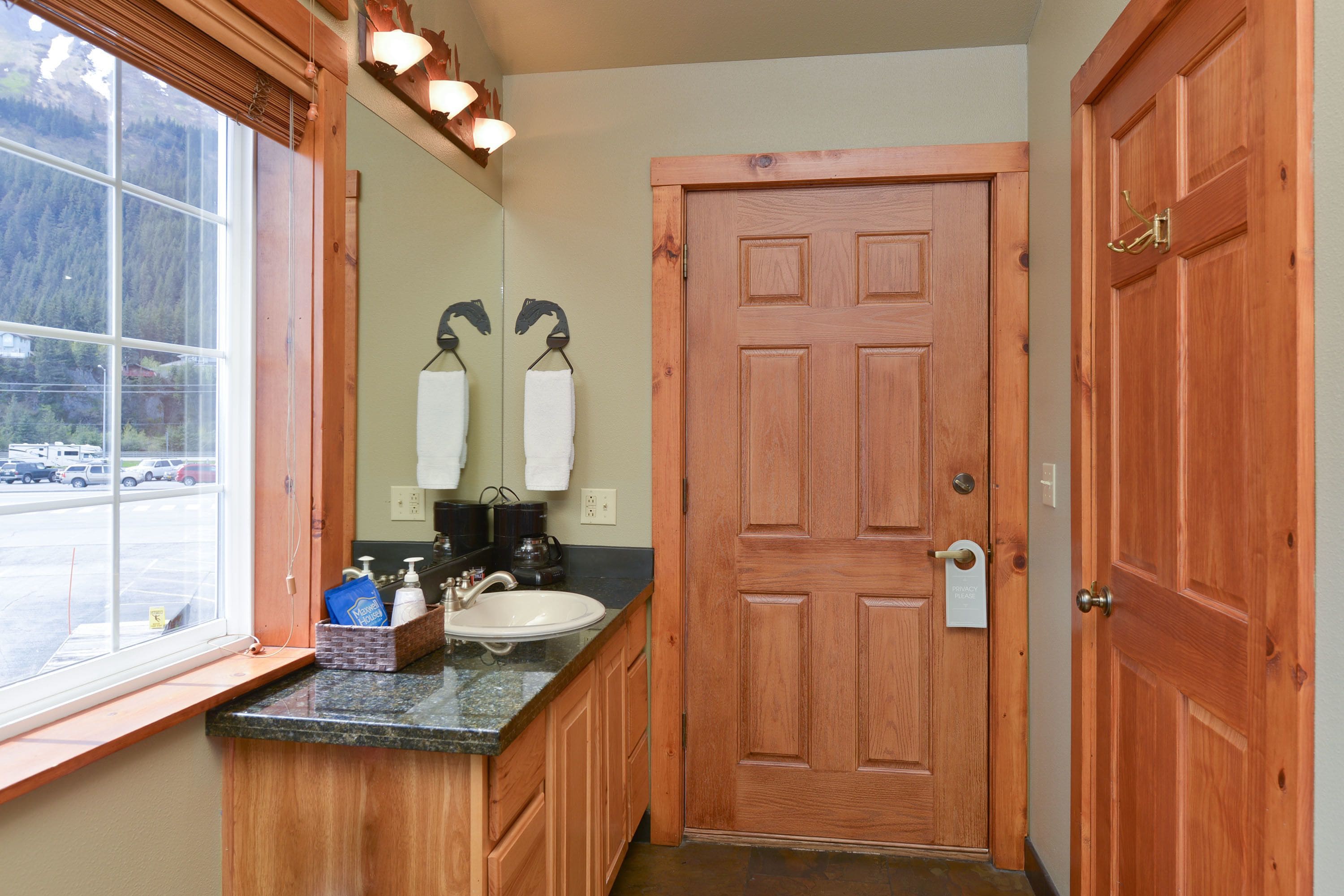 A bathroom with a wooden door, a sink, and a window with a view of parked cars outside.