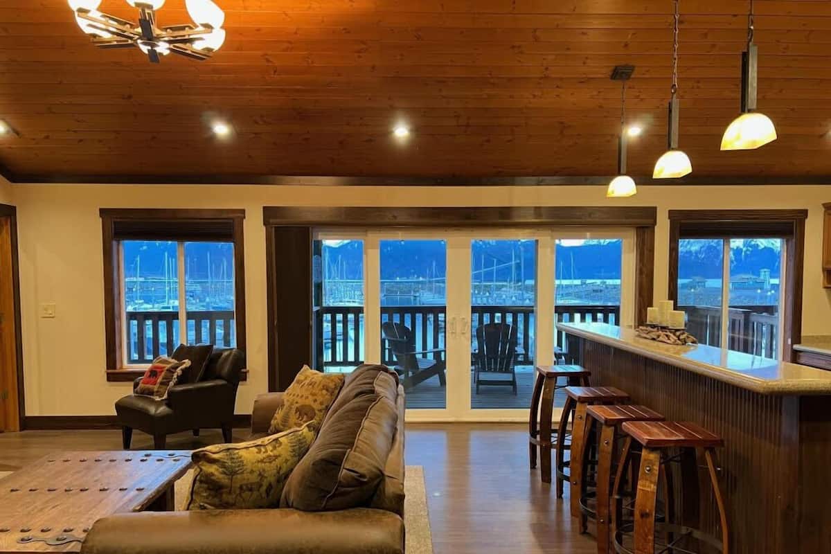 Ein großzügiger Wohnbereich mit Holzboden, einem Ledersofa, einem Holztisch und einer Bar mit Hockern. Große Fenster bieten Blick auf einen Yachthafen.
