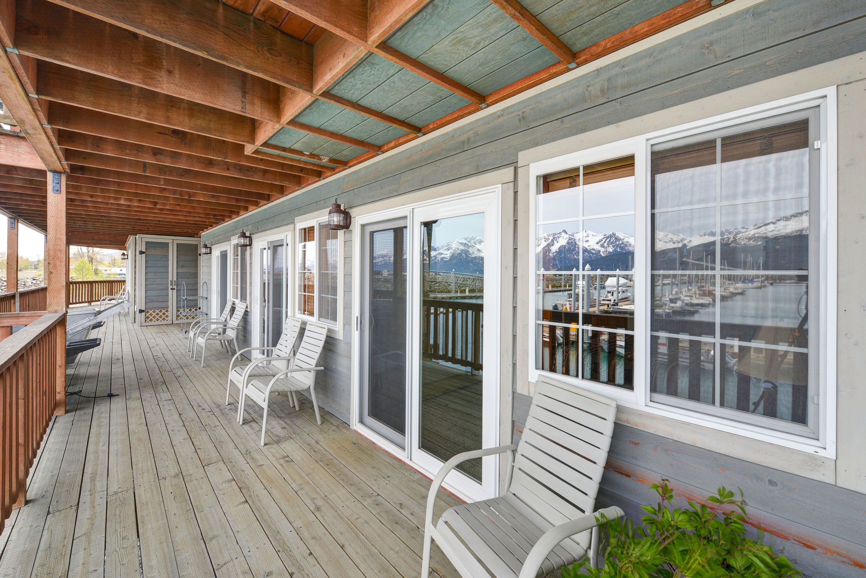 A covered deck with wooden flooring, white chairs, and large windows offering a view of mountains and a marina.